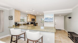 Una cocina con una isla central, dos taburetes de bar, armarios de madera, un microondas y un cuadro de una playa.