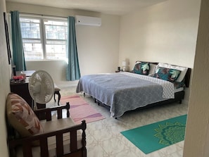 Quarto com cama, escrivaninha, cadeira, ventilador e janela com cortinas.