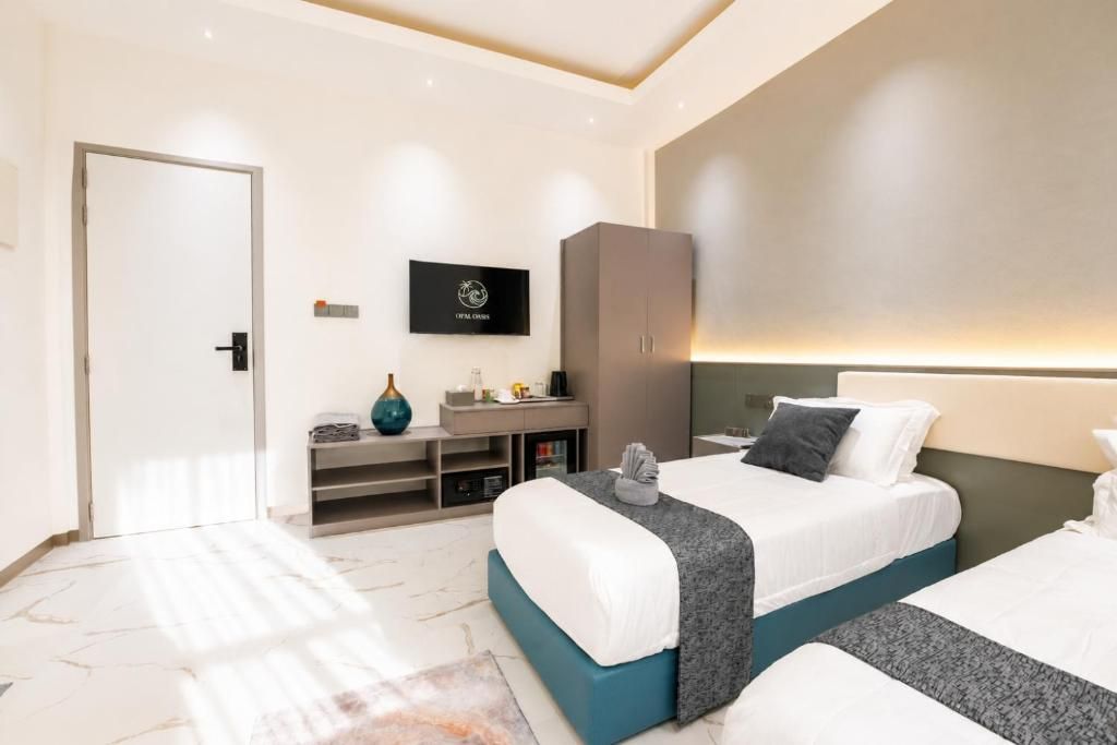 A modern hotel room with two beds, a TV mounted on the wall, a wardrobe, and a small seating area with a vase.