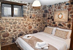 Un dormitorio con pared de piedra, una cama con ropa blanca, una ventana con cortinas de encaje y una obra de arte enmarcada en la pared.