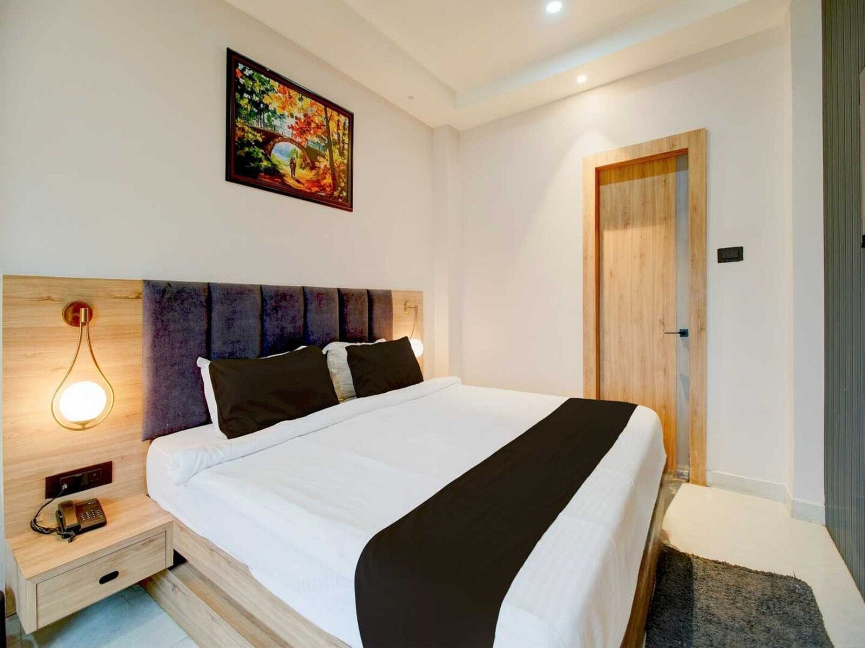 A modern hotel room with a large bed, a nightstand with a lamp, a wooden headboard, a door, and a painting on the wall.