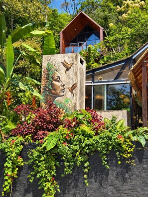 A modern building with a large mural of a face and birds, surrounded by lush greenery and a black brick wall.