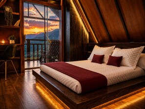 A bedroom with a large bed, a wooden floor, a view of mountains through a glass door, and a balcony with a chair.