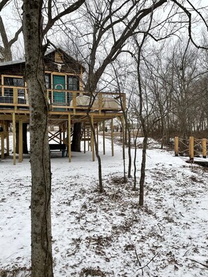 A wooden cabin with a deck, surrounded by trees and snow-covered ground. - Fun & Magical Treehouse (Coweta)