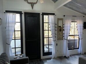 A room with a black door, two windows with white curtains, a ceiling fan, and a small table with a vase.