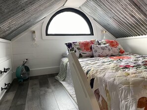 A small, cozy attic bedroom with a sloped ceiling, a single bed with floral bedding, a standing fan, and a window with a curved top. - Fun & Magical Treehouse (Coweta)