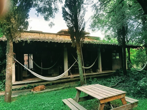A wooden structure with a thatched roof, a hammock, and a picnic table in a lush garden setting.