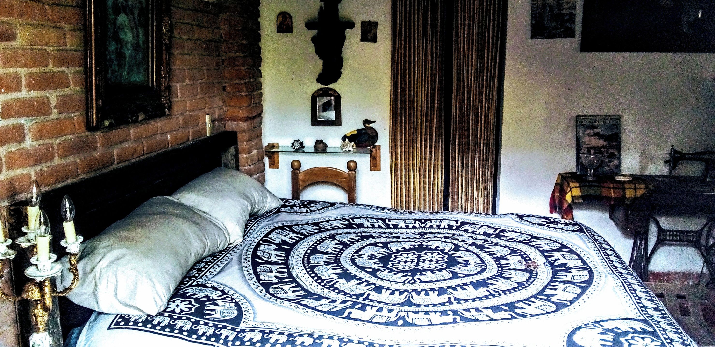 A room with a bed, a wooden headboard, a brick wall, a mirror, a curtain, and a small table with a lamp.