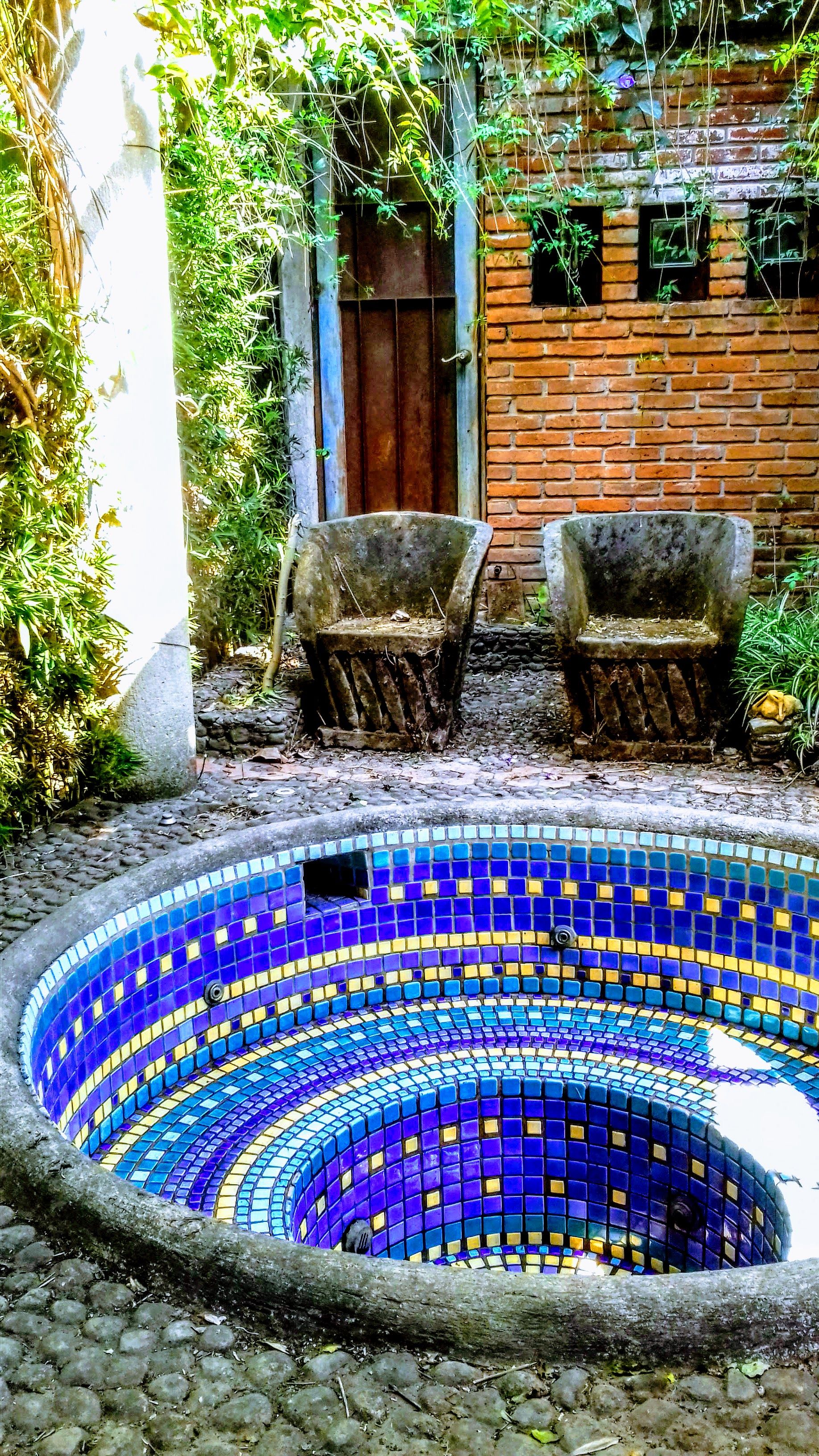 A circular mosaic pool with a brick wall and two chairs in the background.