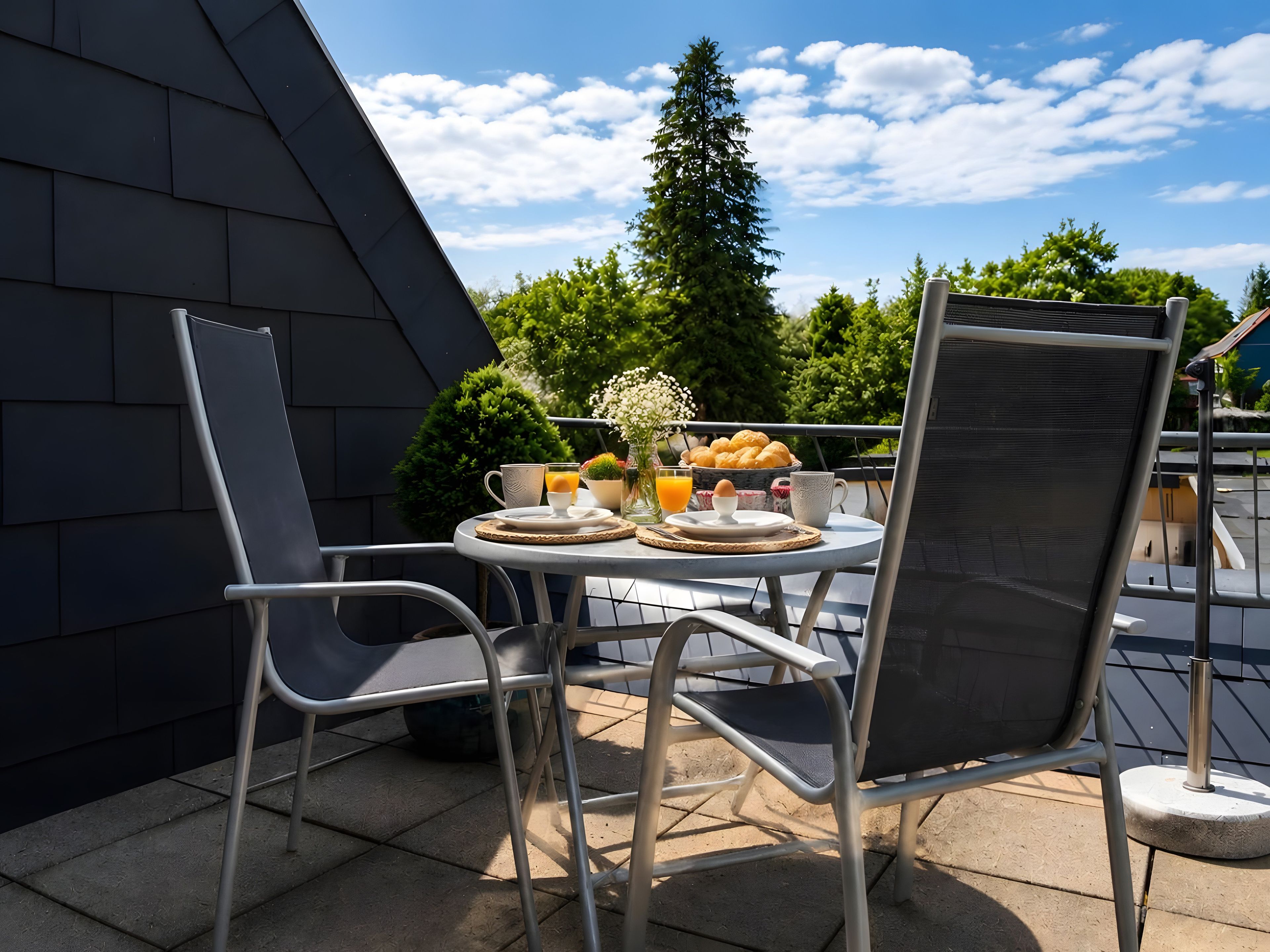 An outdoor dining area with a table set for breakfast, including eggs, bread, and juice, chairs, and a view of trees and a building.