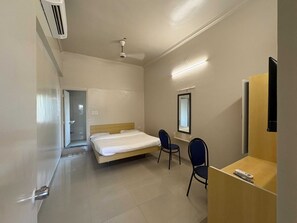 A hotel room with a bed, two chairs, a desk, a mirror, and a ceiling fan. - Deluxe Hotel Saubhagyashanti (Nashik)