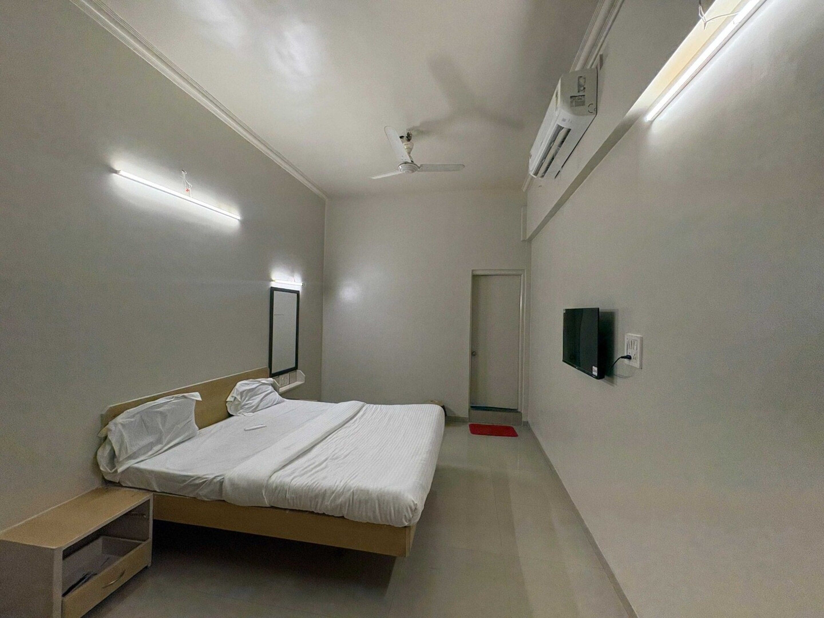 A small, single-bed room with a ceiling fan, a bedside table, a mirror, and a wall-mounted TV.