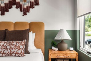 Quarto bem arrumado com cama, travesseiros, mesa de cabeceira com abajur, uma pequena planta e janela com vista para o exterior.
