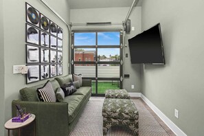 Un salon avec un canapé vert, un pouf à motifs, un téléviseur à écran plat fixé au mur et une fenêtre donnant sur l’extérieur.