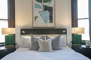 Hotellihuone, jossa on suuri sänky, kaksi yöpöytää, seinälle ripustettu taideteos ja ikkuna, jossa on verhot.