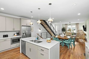 A modern kitchen with white cabinets, stainless steel appliances, and a wooden staircase leading to a dining area with teal chairs and a wooden table. - Wellness Retreat  | 8th + 12th Ave South (Nashville)