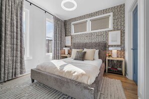 Quarto com cama grande, duas mesas de cabeceira com luminárias, papel de parede estampado e vista do céu pela janela.