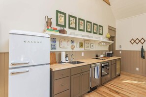 A compact kitchen with a Galanz refrigerator, wooden cabinets, a sink, and a dishwasher. There are framed botanical prints on the wall and a shelf with dishes and a vase. - 7  Cottages at Buckhorn Hollow - Sleeps 28 (McEwen)