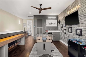 A modern living room with a foosball table, a ceiling fan, and a flat-screen TV mounted on a textured wall. - Luxe Retreats Sleeps 24: Games, Views, Near Dwntwn (Nashville)
