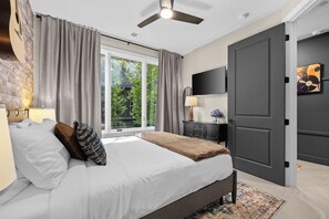 A bedroom with a bed, curtains, a television, a door, and a window with a view of greenery. - Songwriters' Retreat: 4BR, Rooftop Sauna & Bar (Nashville)