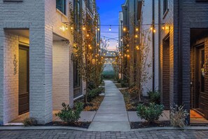 A narrow alleyway with a pathway flanked by buildings with brick facades, string lights, and small plants. - Songwriters' Retreat: 4BR, Rooftop Sauna & Bar (Nashville)