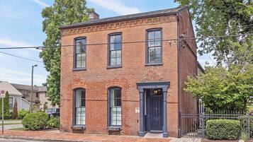 A two-story brick building with a black door and windows, a small porch, and a fenced yard.