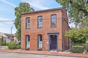 A two-story brick building with a black door and windows, a small porch, and a fenced yard. - The Polk Suite | Germantown Inn | Breakfast (Nashville)