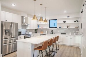 Una cuina moderna amb armaris blancs, electrodomèstics d'acer inoxidable, una illa central amb tamboretes i il·luminació penjant, i un televisor de pantalla plana.