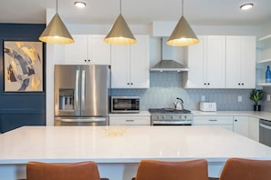 Cozinha moderna com ilha central, eletrodomésticos de aço inoxidável, iluminação pendente e uma parede azul como detalhe.