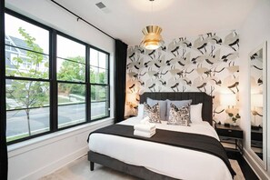 Ein modernes Schlafzimmer mit großem Fenster, einem Bett mit dunklem Kopfteil, gemustertem Wandpapier und Nachttischlampen.