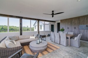 Een moderne woonkamer met een bank, stoelen, een salontafel en een eettafel. Er is een plafondventilator, grote ramen en uitzicht op de omgeving.