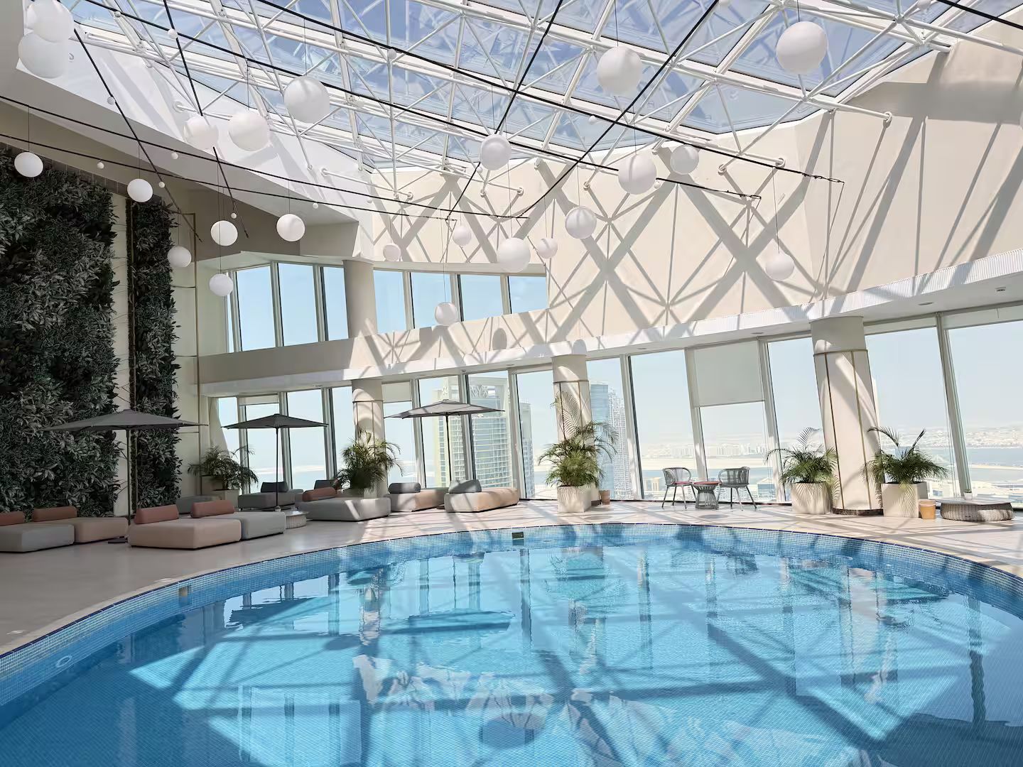A modern indoor swimming pool with a glass ceiling, seating areas, and potted plants.