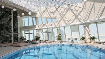 A modern indoor swimming pool with a glass ceiling, seating areas, and potted plants.