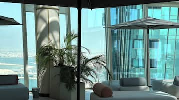 Un área de descanso moderna con una planta grande en maceta, asientos cómodos y vistas de un paisaje urbano a través de amplios ventanales.