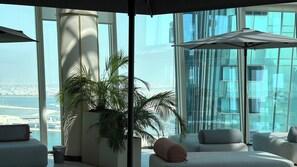 Un área de descanso moderna con una planta grande en maceta, asientos cómodos y vistas de un paisaje urbano a través de amplios ventanales.