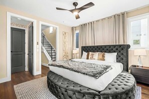A bedroom with a tufted headboard, a large bed with a fluffy blanket, a standing lamp, and a ceiling fan. - Printer's Alley Vibes Rooftop Fire Table + Karaoke (Nashville)
