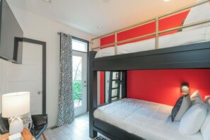 Een stapelbed met een rood hoofdeinde, een bureau met een lamp en een raam met gordijnen.