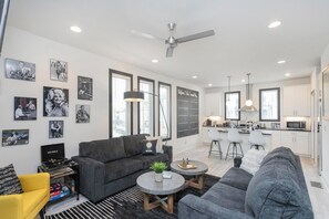 Ein modernes Wohnzimmer mit einem grauen Sofa, einem gelben Sessel und einem Couchtisch. Es gibt eine Küchenzeile mit weißen Schränken und einer Mikrowelle.