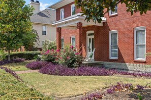 A two-story brick house with a front porch, a well-maintained garden with purple foliage, and a clear sky. - Historic East Nash | 5 Kings, 5 Baths, Walkable (Nashville)