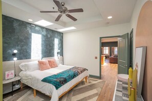 Quarto com cama, ventilador de teto, abajures e vista para outro cômodo.