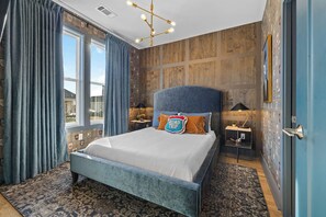 A bedroom with a large bed, a headboard, a nightstand, a lamp, and a wall with various pictures. - 2 Homes | 24 Guests | Rooftop Hot Tub Fire Pit (Nashville)