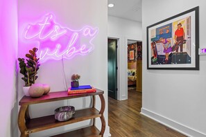Un pasillo con un letrero de neón que dice "it's a vibe", una mesa consola de madera con objetos decorativos y un cuadro en la pared.