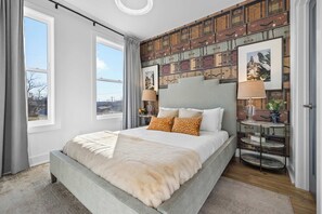 Quarto com cama grande, cabeceira decorativa, parede com padrão e janela com cortinas.