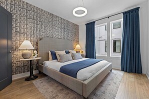 Quarto com cama, luminárias de cabeceira, papel de parede estampado, janela com cortinas e uma luminária de teto redonda.