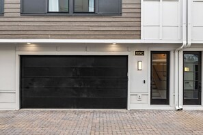 Un bâtiment moderne doté d’un grand portail de garage noir, d’une entrée pavée de briques et d’une façade blanche.