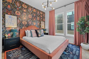 Quarto com papel de parede estampado, cama com várias almofadas, mesa de cabeceira, janela grande com cortinas e um vaso de planta.