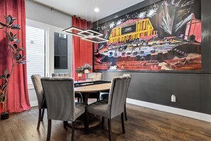 A dining area with a round table, two upholstered chairs, a red curtain, a potted plant, and a large mural of a circus on the wall. - 2 Units Sleeps 24 | Roof Decks Karaoke Murals (Nashville)