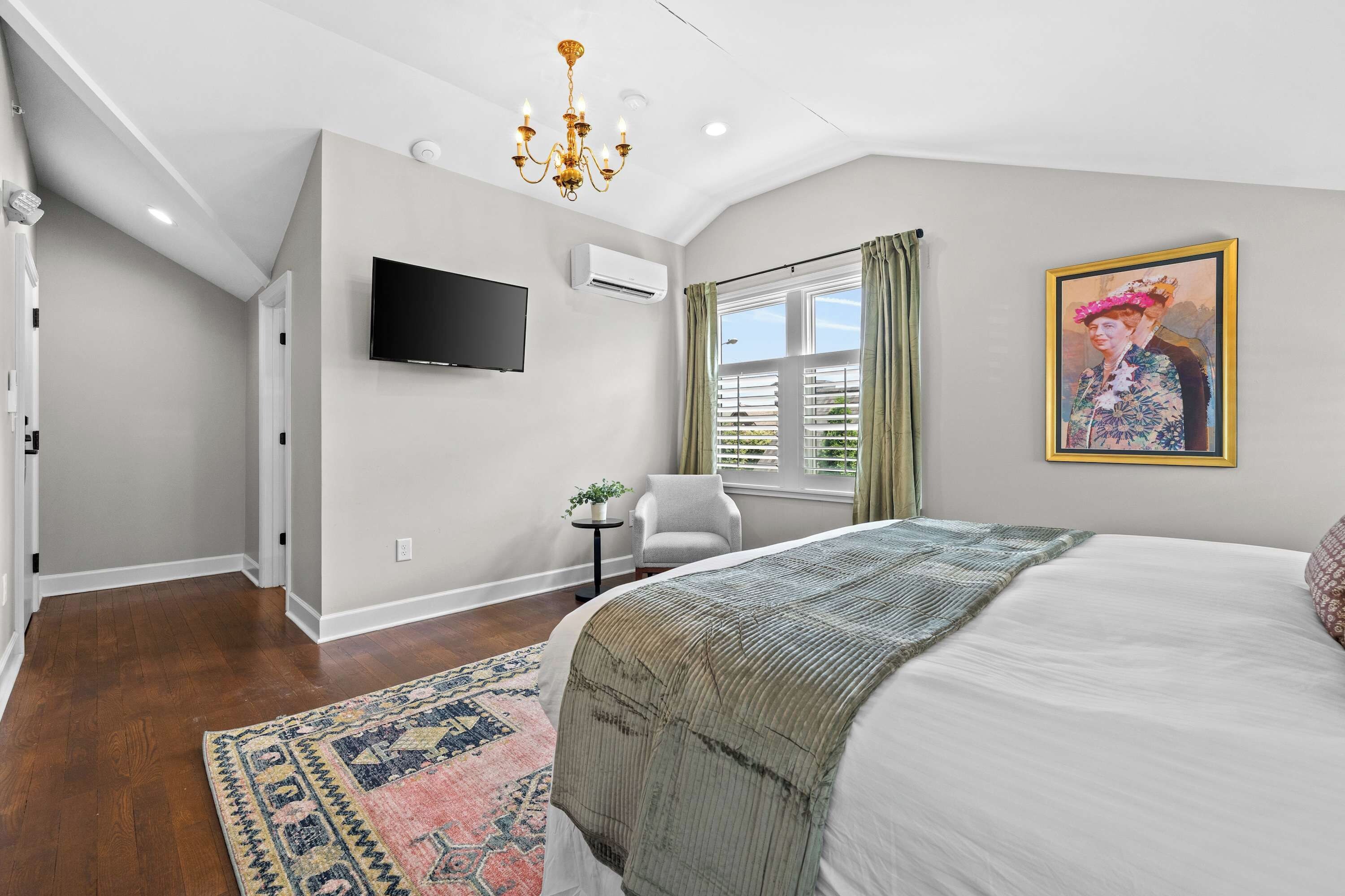 A bedroom with a large bed, a flat-screen TV, an air conditioning unit, a window with curtains, and a painting on the wall.