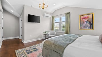 A bedroom with a large bed, a flat-screen TV, an air conditioning unit, a window with curtains, and a painting on the wall.