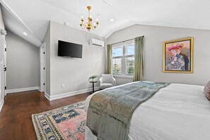 A bedroom with a large bed, a flat-screen TV, an air conditioning unit, a window with curtains, and a painting on the wall.